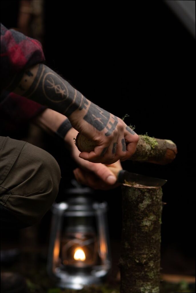 A tattooed arm splitting logs with an axe in a rustic camping setting, lantern lit.