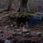 A lone camper sits by a campfire at the base of a tree near a river.